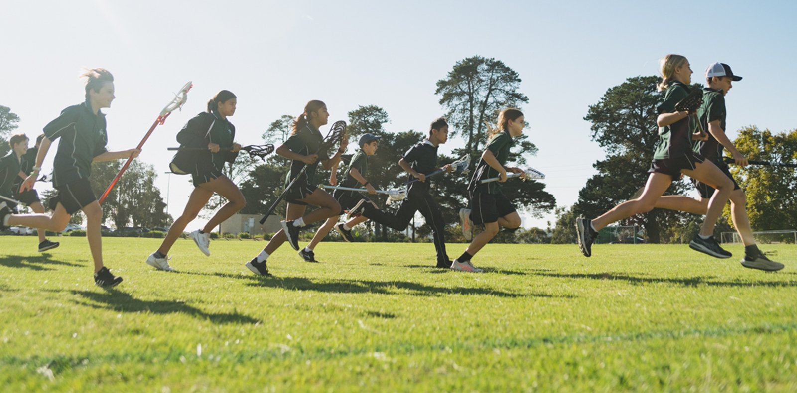 A group of students run across a field together with lacrosse sticks.