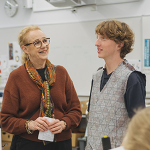 A student and teacher stand together in the classroom having a discussion, while the student wears their textiles creation.