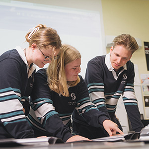 Three students stand gathered around a desk looking at a workbook.